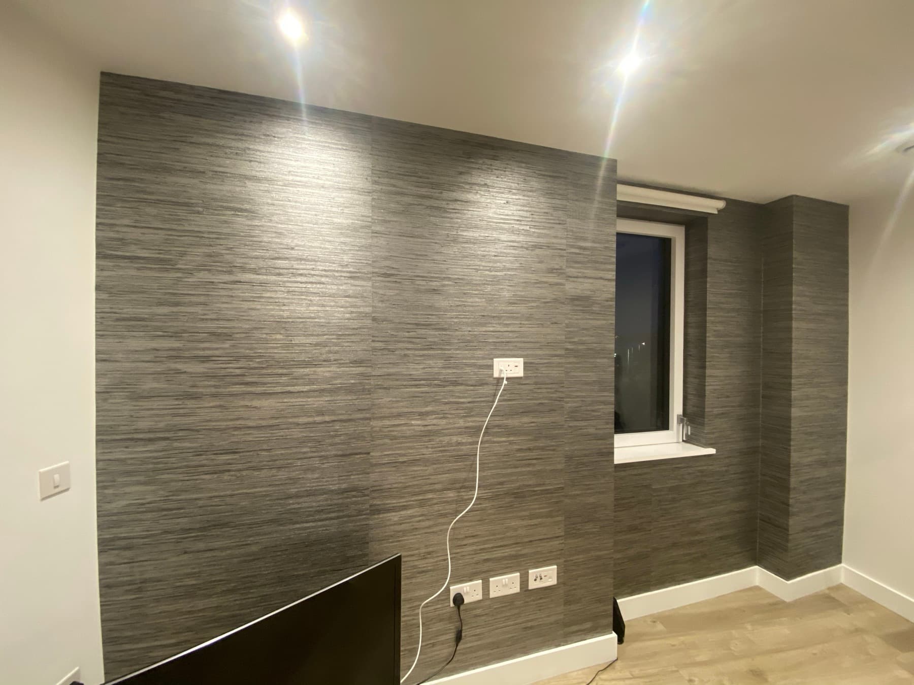 Grey grasscloth textured wallpaper hung across a living room wall and returned into the window reveal